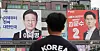 A man wearing a ″Korea″ shirt stands in front of posters for Lee Jae-myung, left, and Kim Moon-soo