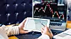 A person holds a tablet showing a financial chart and points at it with a pen. A laptop in the background displays stock market graphs, while printed financial documents rest on the desk. [SHUTTERSTOCK]