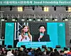Outdoor festival stage with two hosts on large screens and a crowd watching in Seoul.