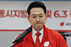 Jang Dong-hyeok speaks at a podium in a red jacket with a microphone and election signage behind him.