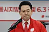 Jang Dong-hyeok speaks at a podium in a red jacket with a microphone and election signage behind him.