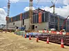 Multi-story building under construction with cranes, cones, fencing, and materials on a dirt site.
