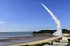 A missile launches from a coastal defense site, trailing white smoke over the sea.