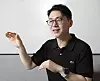 Man in glasses wearing a black polo shirt gestures while seated indoors.