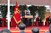 Kim Jong Un stands at a podium on an outdoor stage with officials seated behind him and a red party flag beside him.