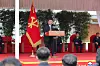 Kim Jong Un stands at a podium on an outdoor stage with officials seated behind him and a red party flag beside him.