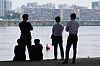 Four people stand and sit by the Han River, facing buildings across the water in Seoul.