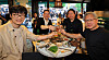 Four men sit at a round table with Korean barbecue dishes and raise drinks inside a restaurant.