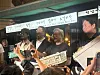 Customers stand in front of a Korean street food stall with menu signs and trays of food.