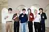 BoyNextDoor standing side by side indoors in front of arched wall decor, making hand gestures.