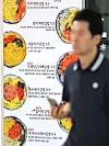 A blurred man passes in front of a Seoul restaurant menu board showing photos of gimbap dishes and prices.