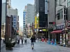 People walking on a wet city street with storefronts and tall buildings on both sides.
