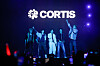 CORTIS band members stand on a lit stage with purple lights and audience phones in the foreground.