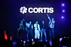 CORTIS band members stand on a lit stage with purple lights and audience phones in the foreground.