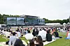 Large crowd seated on grass facing an outdoor concert stage at a festival.