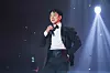 A singer in a black suit holds a microphone on stage beneath dramatic spotlights.