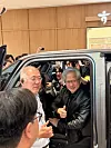 People crowd around a vehicle indoors while two men inside pose with thumbs up.