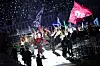 Performers wave flags on a brightly lit stage with a crowd in the stands behind them.