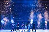 A group of performers stands on a stage under bright fireworks and sparks in a stadium.