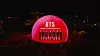 Red illuminated dome with BTS lettering and group silhouettes at a nighttime city event.
