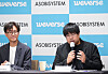 Two men seated at a press conference table in front of a Weverse and Asobisystem logo wall.