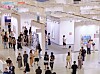 Crowded art gallery interior with visitors walking past framed works on white walls.