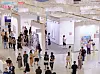 Crowded art gallery interior with visitors walking past framed works on white walls.