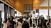 Crowded indoor exhibition hall with booths, overhead lighting, and people walking through the aisle.