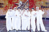 A group of performers in white stands together on a stage with a vivid orange backdrop.
