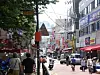 Crowded city street with pedestrians, scooters, shop signs and buildings lining both sides.