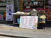 Sidewalk food stall with covered serving trays and a large menu board in front of a shop.