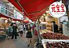 People walking through a covered market street with red awnings and food stalls displaying roasted items.