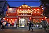 Red-lit restaurant storefront with pedestrians walking past on a street at dusk.