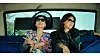 Two passengers in sunglasses sit in a bus, one making a peace sign.