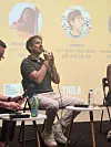 Man speaking into a microphone during a panel event with name profiles on a yellow backdrop.
