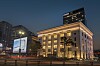 A lit corner building at dusk with light trails from passing traffic and nearby high-rises.