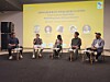 Five panelists seated on stage during a conference discussion with a yellow presentation backdrop.