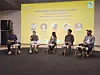Five panelists seated on stage during a conference discussion with a yellow presentation backdrop.