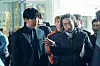 Two men in dark coats stand in front of a group inside a bright modern building.