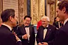 Four men in tuxedos and suits stand conversing at a formal dinner inside an ornate palace room.