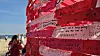 Red and white ribbons with handwritten messages tied to a fence by the beach.