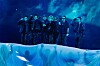 A group of performers standing on a blue-lit stage behind draped fabric.