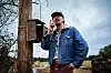 Man in a denim jacket talks into a handheld radio next to a utility pole outdoors.