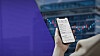 Hand holding a smartphone displaying an omnibus account screen with trading charts behind it.