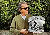 Man wearing sunglasses holds a decorated white guitar in front of a leafy hedge.