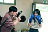 Two actors wearing boxing gloves spar in a bright training room.