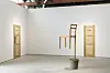 Sparse gallery room with a chair mounted on a pole, a small table, and wooden panels on white walls.