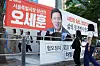 People gathered behind barriers next to a large red and white political campaign banner with a portrait.