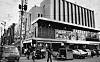 People walk near a large corner building at a busy city intersection with buses and cars.