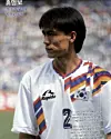 Soccer player in a white South Korea jersey with colorful stripes and number 2, standing in front of a blurred crowd.